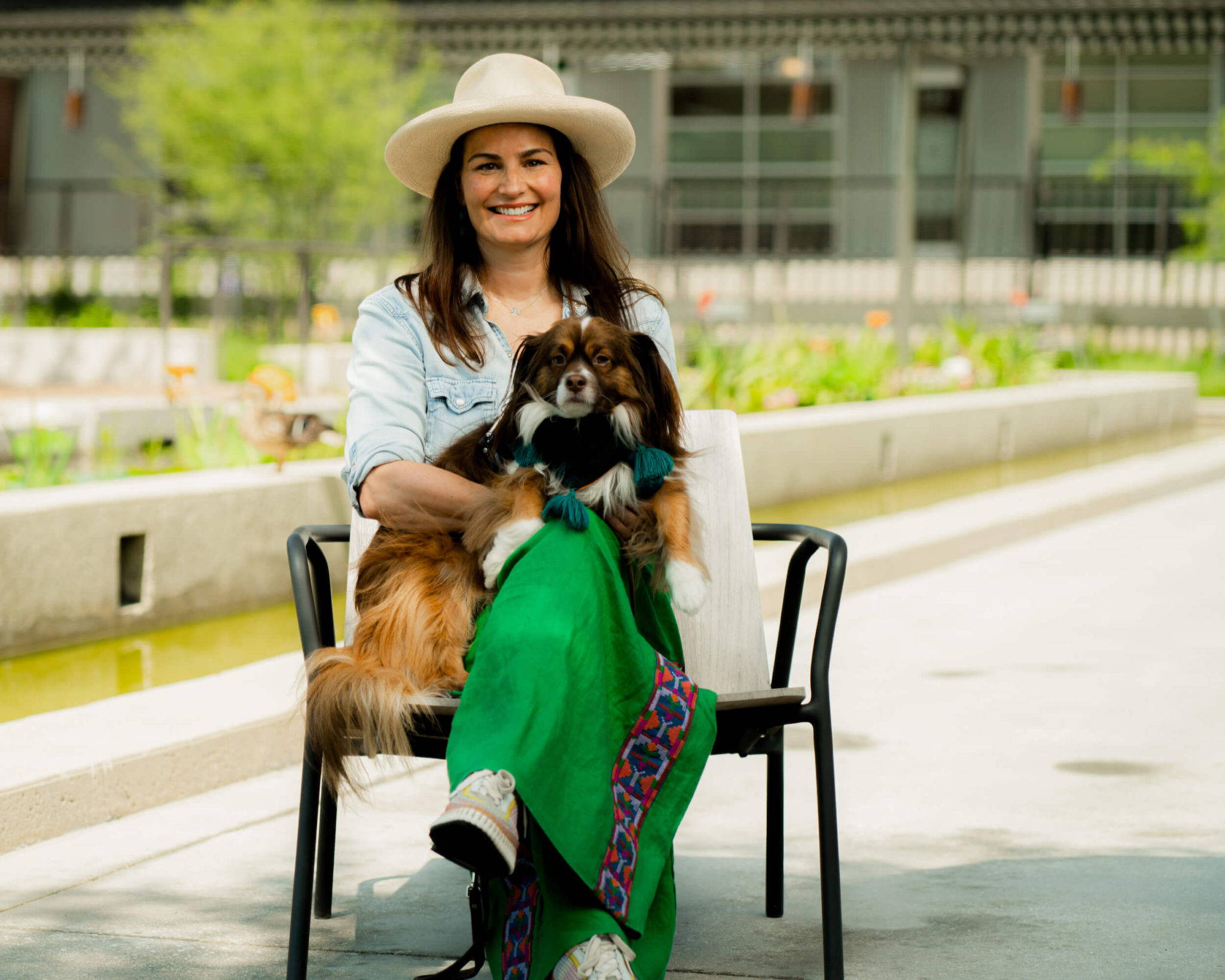 woman wearing cream hat, light denim shirt, and green pants sitting with long haired brown and white dog in her lap outside in front of fountain