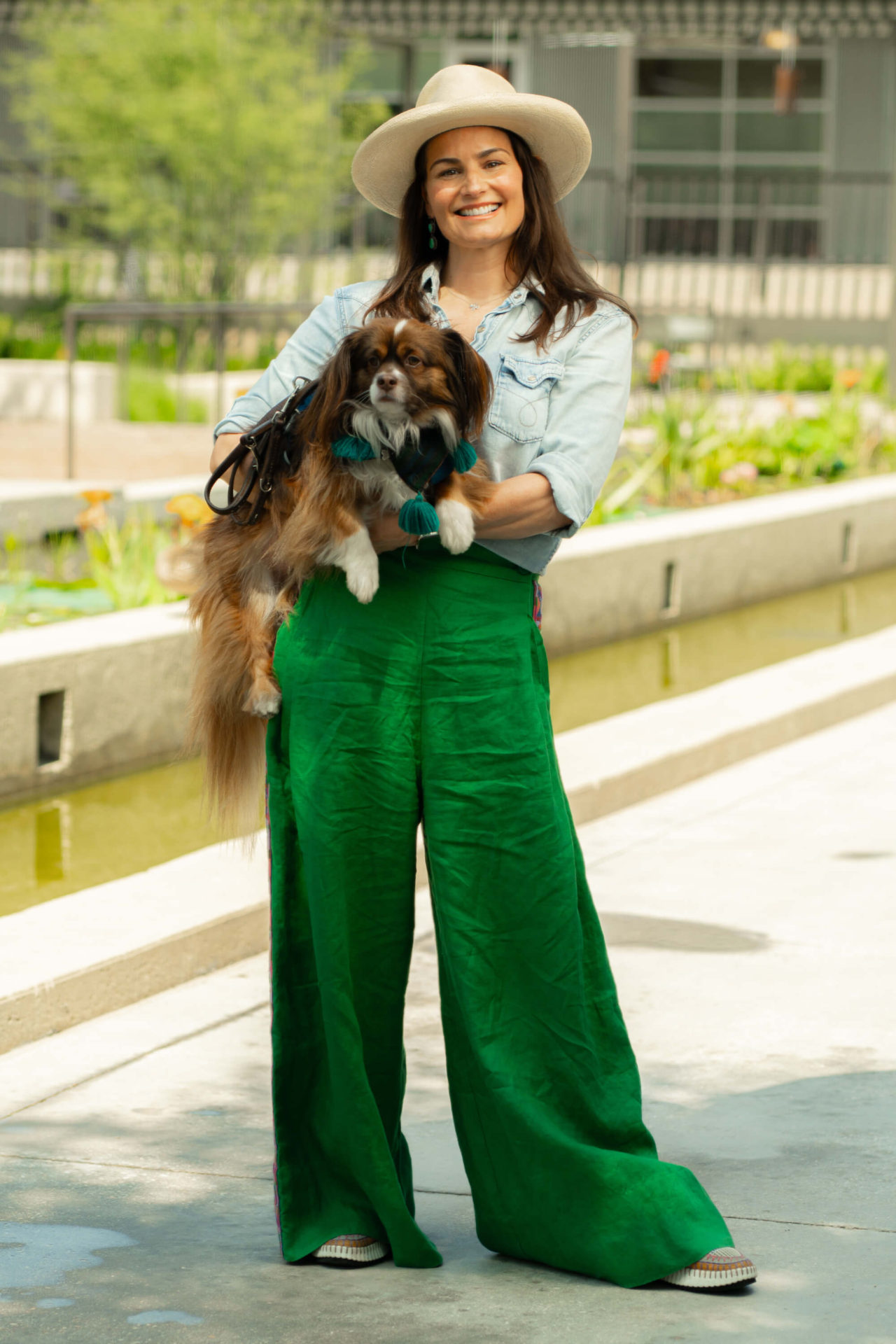 woman with green pants, denim shirt, and cream colored hat standing up and holding chihuahua pekingese dog in front of fountain